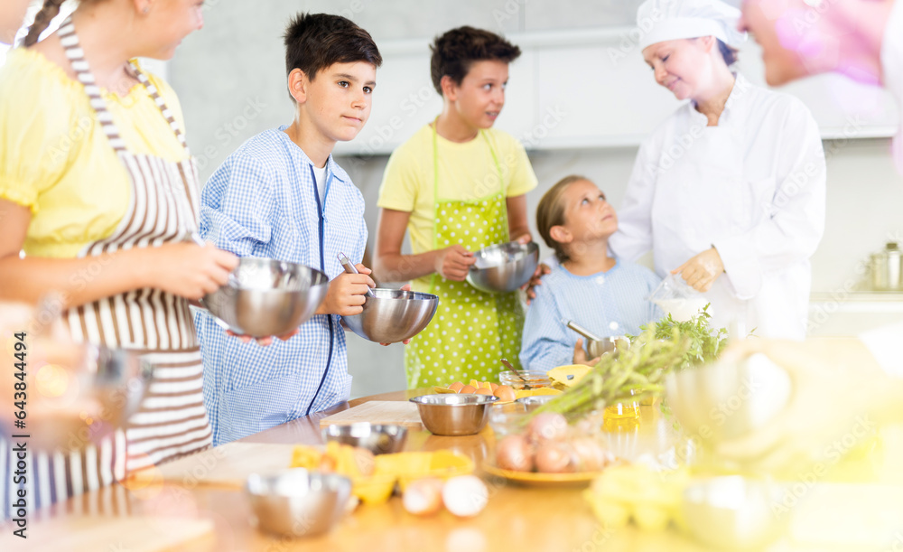 Interested tween boy taking part in cooking classes for children, mixing sauce in bowl with whisk, carefully listening to advice of professional chef instructor