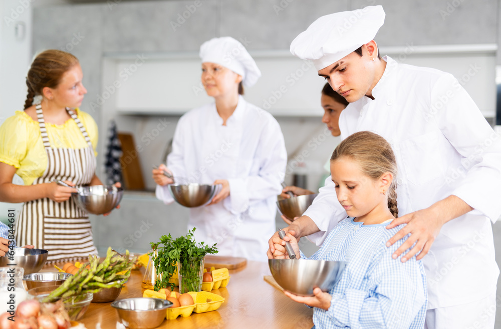 In restaurant kitchen during children cooking class, male chef help small girl thoroughly stir homogeneous liquid dough with whisk. In background, blurry participants are standing near table with food