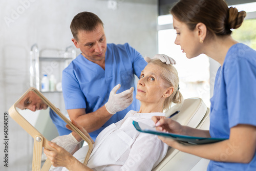 Interested elderly woman sitting on examination couch and looking at mirror while professional cosmetologist with female assistant planning and explaining future rejuvenating facial procedure