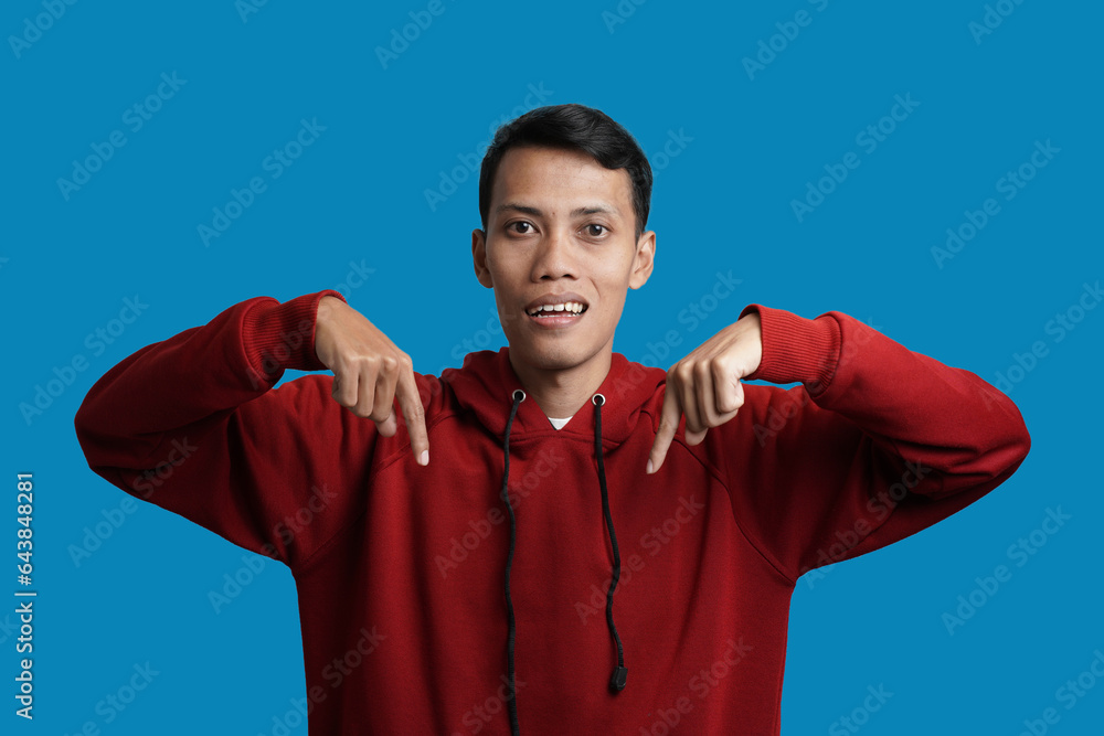 Handsome cheerful young Asian man wearing red hoodie pointing at copy space place, showing place for your advertisement isolated over blue background. Handsome cheerful young Asian man wearing red hoodie pointing at copy space place, showing place for your advertisement isolated over blue background.