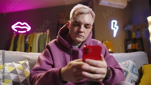 Fair-haired guy with phone on sofa in living room-studio neon lights in background. Expression of negative emotions anger disappointment frustration. Writes messages in anger. Virtual modern lifestyle