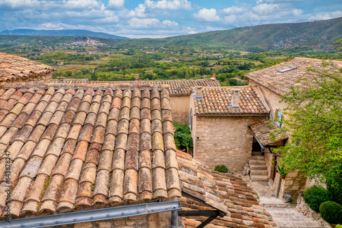 In historic Lacoste in Provence, France, traditional old stone houses with tiled roofs overlook the lush French countryside, with the village of Bonnieux visible in the distance.