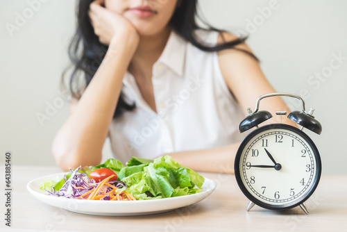 Intermittent fasting with clock, asian young woman, girl dieting, waiting time to eat ketogenic low carb, green vegetable salad on plate. Eat food healthy first meal on brunch, lunch on table at home