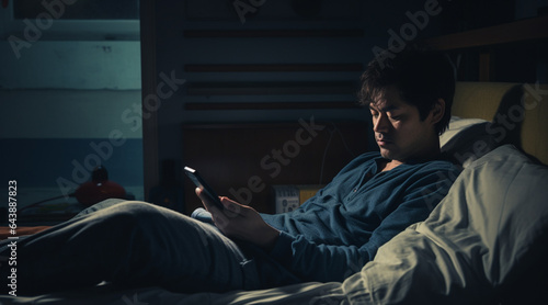 暗い部屋でベッドに寝ながらスマホを触る男性 © Hanako ITO