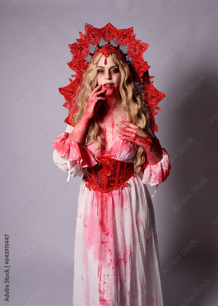 Fototapeta premium Close up portrait of scary vampire zombie queen, wearing elegant halloween fantasy costume with crown and bloody red paint splatter. Isolated on studio background with gestural hands reaching out tow