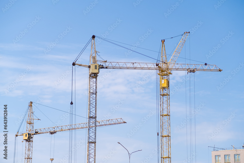 A crane and a building under construction against a blue sky background. Builders work on large construction sites, and there are many cranes working in the field of new construction. A crane and a building under construction against a blue sky background. Builders work on large construction sites, and there are many cranes working in the field of new construction.