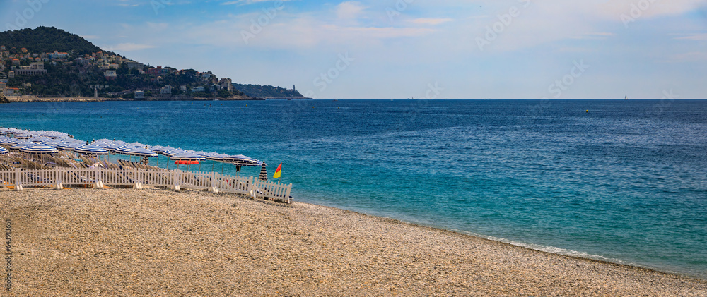 Fototapeta premium Pebble beach along Promenade des Anglais with beach umbrellas and chairs with the turquoise water of the Mediterranean Sea in Nice, Cote d'Azur France
