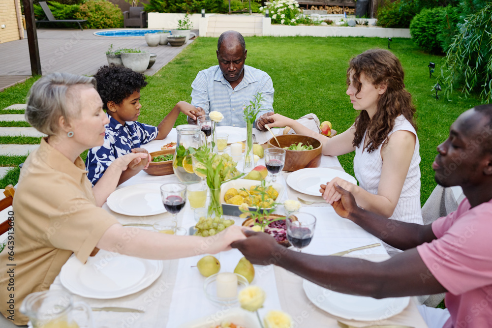 Large intercultural family of three generations holding by hands during pray while sitting by table served with homemade food and drinks Large intercultural family of three generations holding by hands during pray while sitting by table served with homemade food and drinks