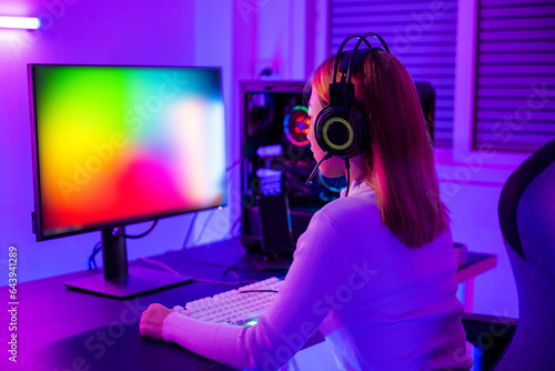 Asian professional gamer playing online video game on desktop computer PC have colorful neon LED lights dark room, young woman in gaming headphones using computer for playing game at home, Back view © sorapop