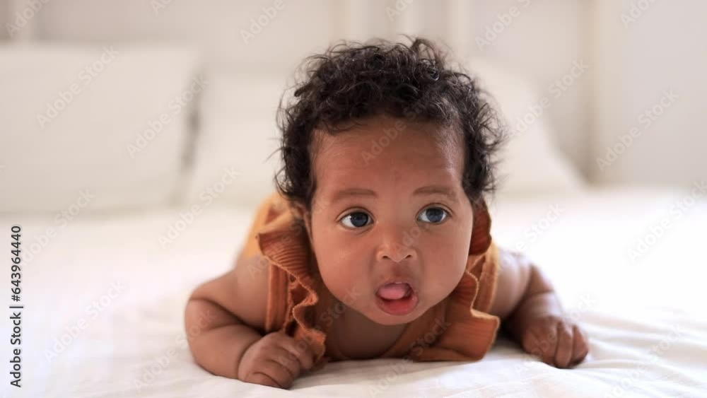 a close-up portrait of a small African-American baby girl in an orange bodysuit on the bed at home, a funny six-month-old black newborn baby lies on the back and smiles. View from above a close-up portrait of a small African-American baby girl in an orange bodysuit on the bed at home, a funny six-month-old black newborn baby lies on the back and smiles. View from above
