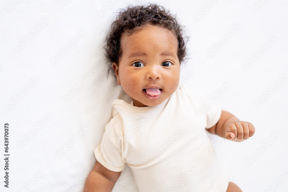 close-up portrait of a small African American baby girl in a white bodysuit on a cotton bed at home, funny six-month-old smiling joyful black newborn baby shows tongue close-up portrait of a small African American baby girl in a white bodysuit on a cotton bed at home, funny six-month-old smiling joyful black newborn baby shows tongue