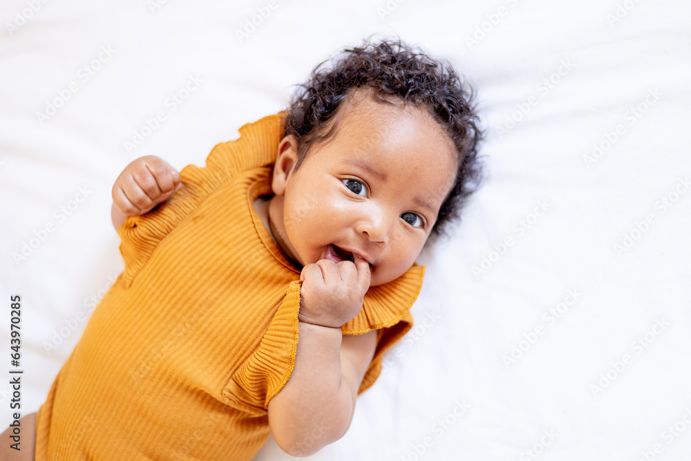 a close-up portrait of a small African-American baby girl in an orange bodysuit on the bed at home, a funny six-month-old black newborn baby lies on the back and smiles. View from above a close-up portrait of a small African-American baby girl in an orange bodysuit on the bed at home, a funny six-month-old black newborn baby lies on the back and smiles. View from above