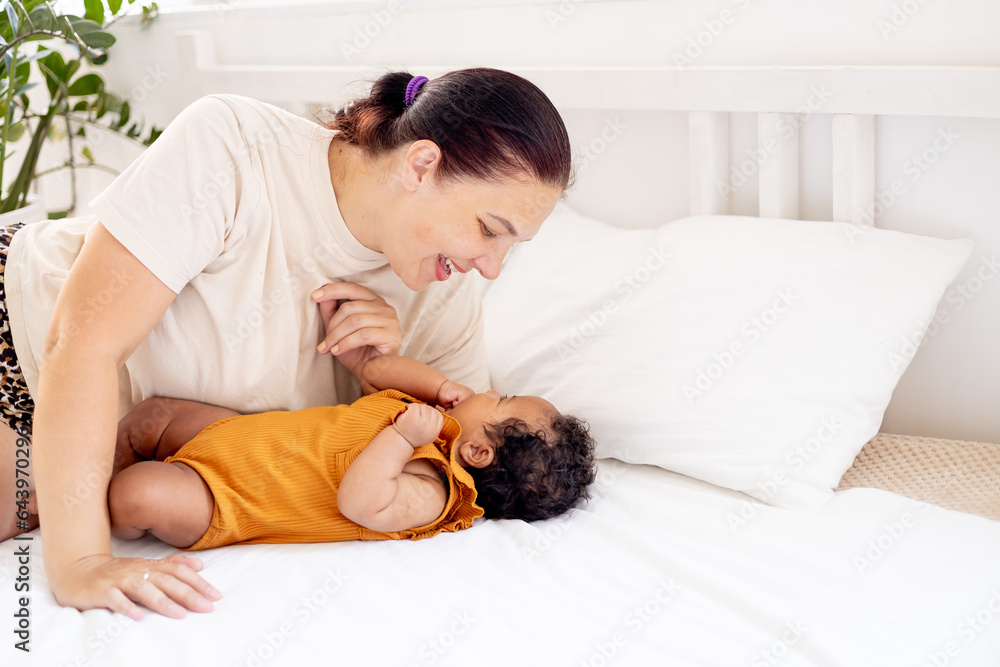 a little African-American baby girl in an orange bodysuit on the bed at home, a funny six-month-old black newborn baby hugs a stuffed rabbit and smiles. View from above a little African-American baby girl in an orange bodysuit on the bed at home, a funny six-month-old black newborn baby hugs a stuffed rabbit and smiles. View from above