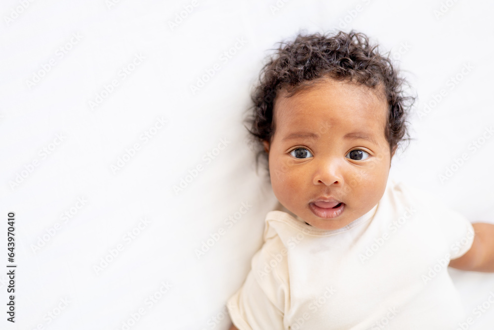 close-up portrait of a small African-American baby girl in a white bodysuit on a cotton bed at home, a six-month-old smiling joyful black baby close-up portrait of a small African-American baby girl in a white bodysuit on a cotton bed at home, a six-month-old smiling joyful black baby