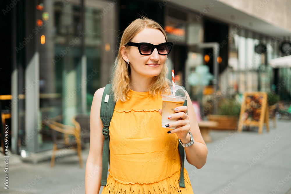 Fototapeta premium Beautiful fashionable young woman in sunglasses with backpack standing on city street. Urban lifestyle concept. Attractive female drinking a ice coffee in plastic cup.
