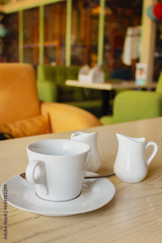 Breakfast service. Coffee, milk and sugar in white utensil. Coffee mug and milk jug on the table in cafe. Cafe interior. White tableware. Morning coffee concept. Breakfast service. Coffee, milk and sugar in white utensil. Coffee mug and milk jug on the table in cafe. Cafe interior. White tableware. Morning coffee concept.