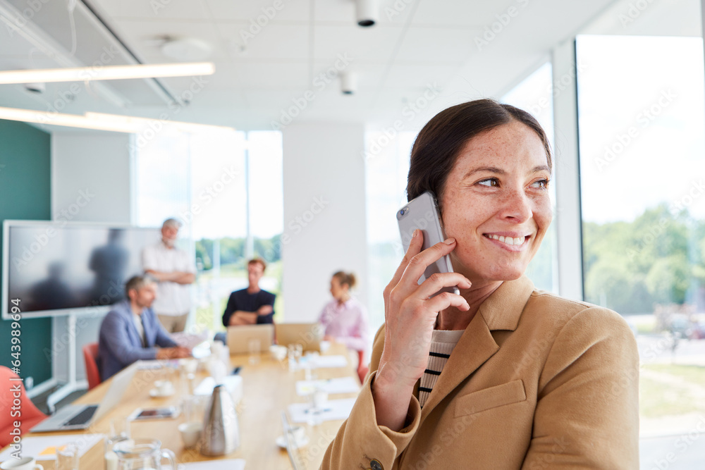 Happy female professional talking on smart phone with team discussing in conference room at workplace Happy female professional talking on smart phone with team discussing in conference room at workplace