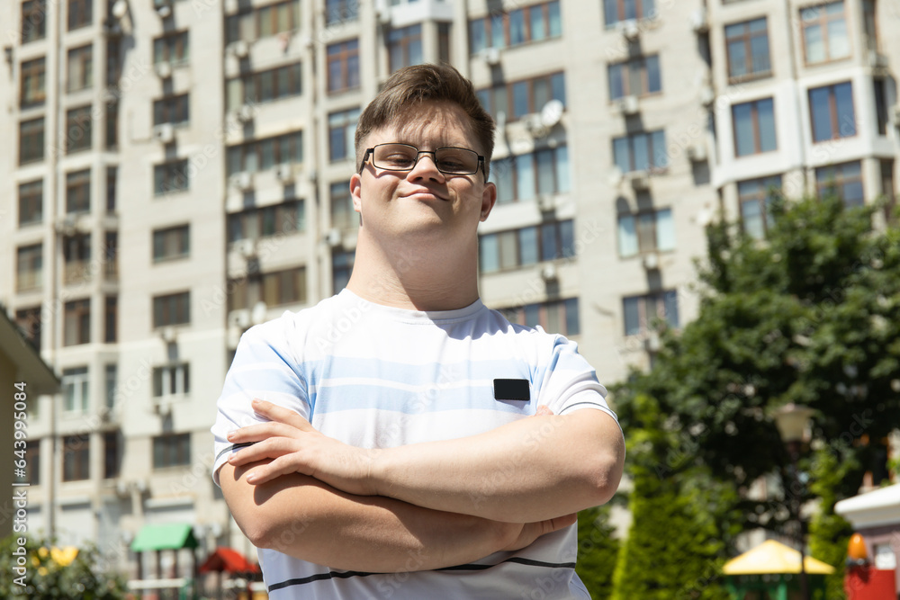 A smiling young man with Down syndrome in glasses poses against the background of the city in the summer A smiling young man with Down syndrome in glasses poses against the background of the city in the summer