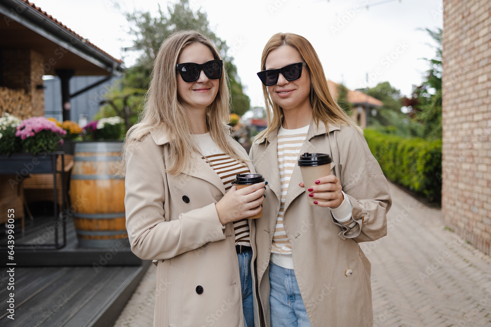 Two stylish girlfriends in a beige trench coat walking around a European city with coffee to go in a paper cup in black sunglasses. Friendship, sisters, blondes, blond hair, tourists in the city