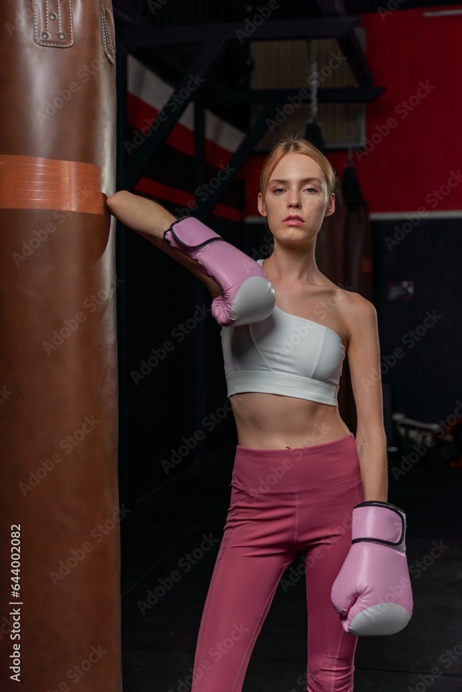 Beautiful and sexy blonde woman wearing pink gloves strikes a confident pose beside a punching bag in a boxing gym, exuding strength and style