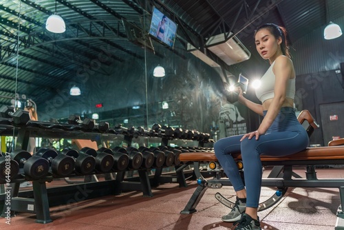 Determined young Asian woman strengthens her arm with single-arm dumbbell rows on the bench press, showing her dedication to building muscle at the gym © asean studio