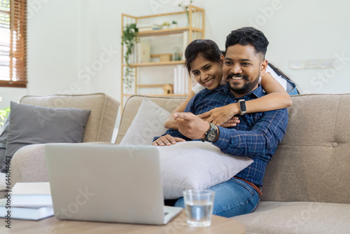 Happy loving family Indian couple hugging on couch at home and focused on laptop screen watching romantic movie