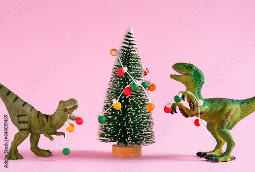 Happy cute green dinosaurs decorate Christmas tree with retro bulb garland. Christmas and new year greeting card on pastel pink background. © dvulikaia