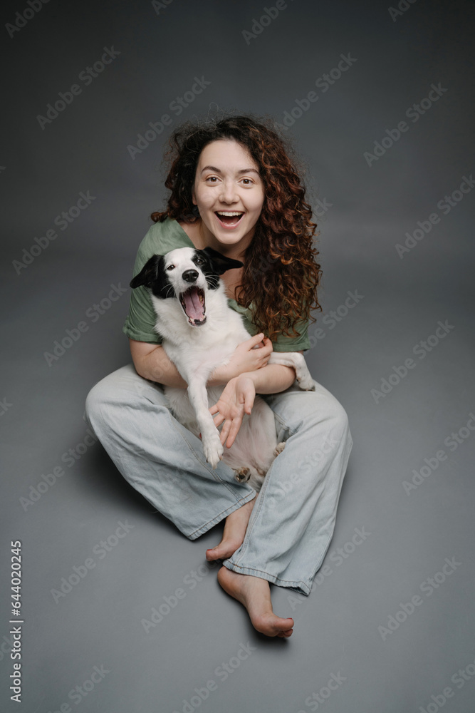 Happy woman hugging her border collie dog happy laughing and looking at the camera. Grey background studio shot. Sitting on floor in blue jeans barefoot