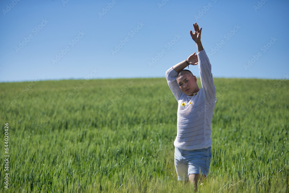 Obraz premium Young, South American, non-binary person, heavily makeup, posing in a white sweater with natural daisies, in the middle of a green wheat field, with one arm raised. Concept queen, lgbtq+, pride, queer