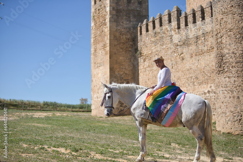 Non-binary person, young and South American, very makeup, mounted on a white horse, smiling and happy, with a gay pride flag on the rump, next to an old medieval castle. Concept queen, lgbtq+, pride. © Manuel