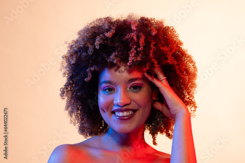 Smiling biracial woman with curly hair in eyeshadow and lipstick touching head in red and blue light