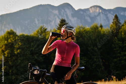 Fototapeta Naklejka Na Ścianę i Meble -  Cyclist silhouette. Female cyclist in cycling clothes and a helmet drinks water from a sports bottle at sunset.