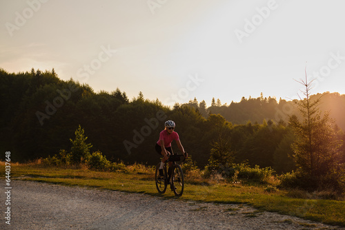 A fit female cyclist, wearing a cycling kit and helmet, is riding a gravel bike on a gravel road at sunset. Sports motivation image.