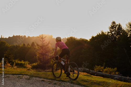 Fototapeta Naklejka Na Ścianę i Meble -  A fit female cyclist, wearing a cycling kit and helmet, is riding a gravel bike on a gravel road at sunset. Sports motivation image.
