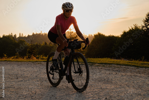 A female cyclist, wearing a pink cycling jersey and a white helmet, is riding a bicycle on a gravel road at sunset.Woman cycling in the nature. Active lifestyle concept. Cycling gravel adventure.