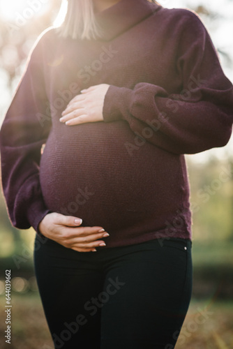 Pregnant woman hugs round belly at sunset closeup. Pregnant girl stands in background of golden leaves in forest. Female walk in autumn park. Pregnancy, fresh air, the concept of relaxing in nature.