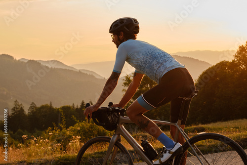 Man cyclist wearing blue cycling jersey. He is riding a gravel bike on a gravel road at sunset with a view of the mountains.Empty mountain road.Cycling gravel adventure in Romania.Bucegi Natural Park photo