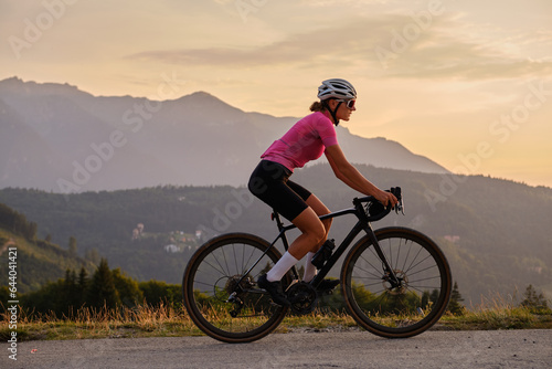 Fototapeta Naklejka Na Ścianę i Meble -  Woman cyclist is riding a gravel bike on a gravel road  at sunset with a view of the mountains.Empty mountain road. Cycling gravel adventure in Romania. Bucegi Natural Park.