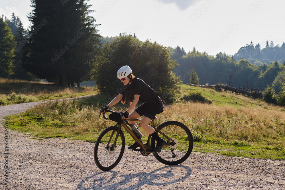 Woman cyclist, dressed in a black cycling kit and a white helmet,is riding a gravel bicycle on a gravel road with beautiful Romanian mountains in the background.Cycling adventure. Bucegi Natural Park.