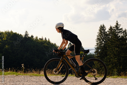 Fototapeta Naklejka Na Ścianę i Meble -  Woman cyclist wearing black cycling kit. She is riding a gravel bike on a gravel road at sunset with a view of the mountains.Empty mountain road.Cycling gravel adventure in Romania.Bucegi Natural Park
