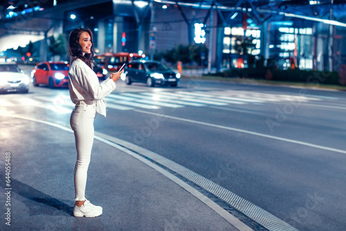 Young woman standing on the street in the night holding smartphone, waiting for taxi, full length, copy space © Prostock-studio