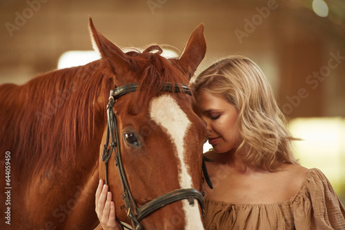 Standing and embracing the animal. Beautiful young woman is with horse indoors