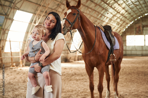 Holding kid in hands. Gorgeous woman with her little baby son is with horse indoors © standret