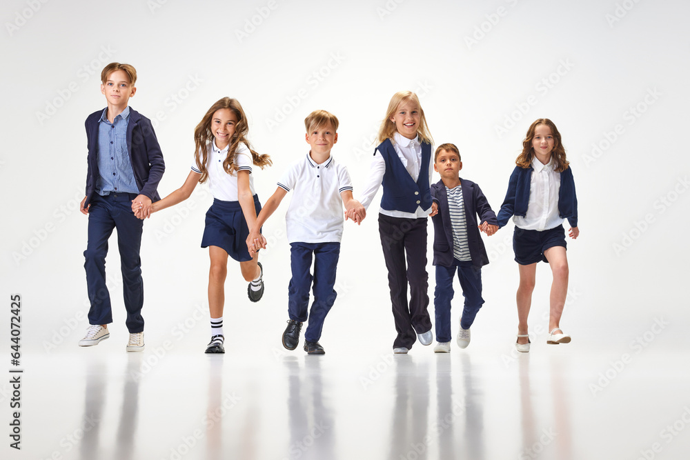 Obraz premium Group of stylish children, boys and girls, pupils in school uniform running against white studio background. Classmates. Concept of childhood, school, education, fashion, style. Copy space for ad