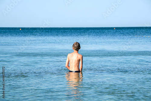 summer, a tanned teenage boy stands with his back to the camera in the sea up to his knees. concept of children and sea, summer holidays and vacations