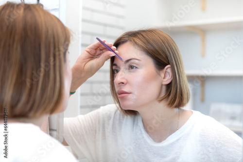 Young woman depilates her face with eyebrow tweezers in front of the mirror. Beauty and self care concept