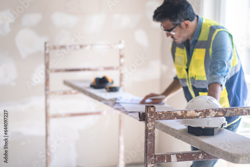 Concentrated specialist in glasses examining construction documentation standing near safety equipment man in vest preparing for reconstruction process in apartment