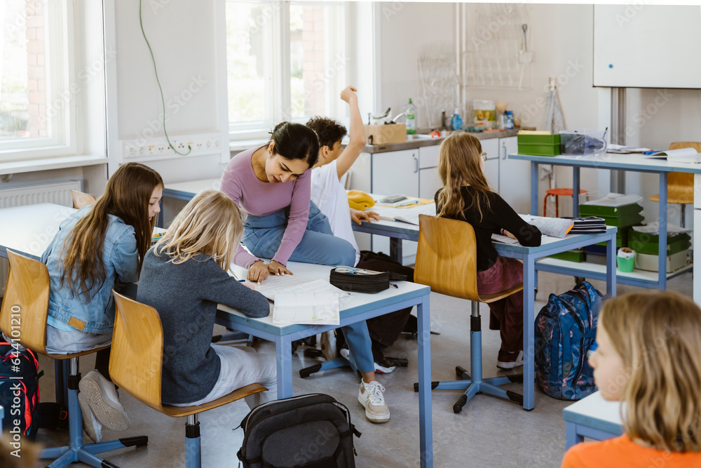 Female teacher assisting schoolgirls while sitting on desk in classroom Female teacher assisting schoolgirls while sitting on desk in classroom