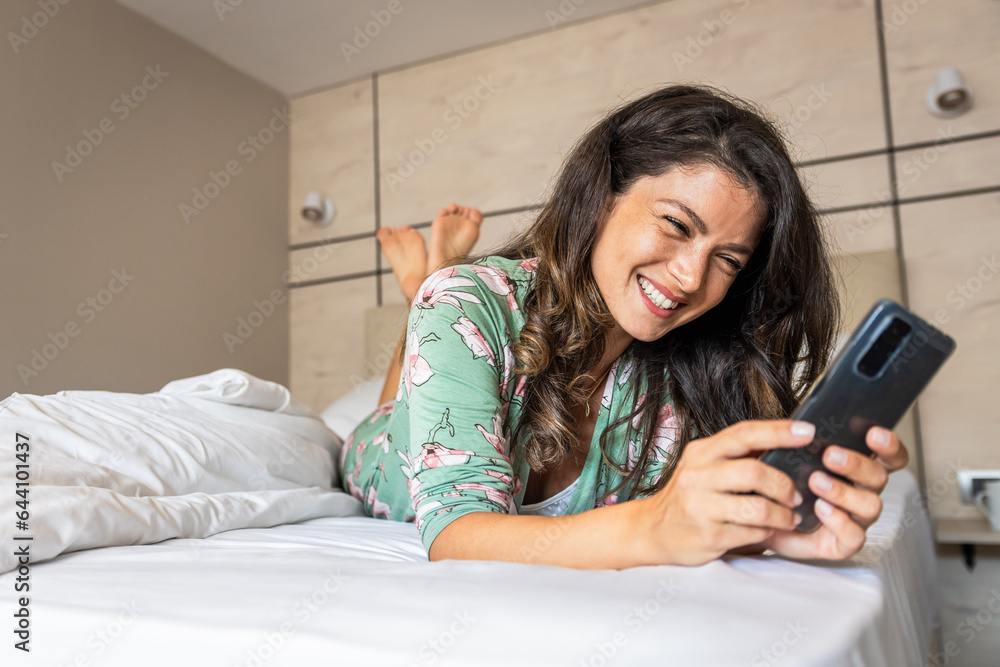 Smiling woman using smart phone while laying down on bed in bedroom in morning at home. Smiling woman using smart phone while laying down on bed in bedroom in morning at home.