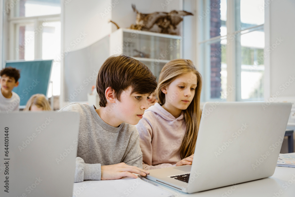 Concentrated boy watching laptop with female friend at desk in classroom Concentrated boy watching laptop with female friend at desk in classroom